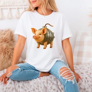 May include: White t-shirt with a cartoon pig design. The pig has a bomb-shaped body, a smiling face, and brown legs. The bomb body is metallic with a lit fuse. The shirt is a casual, unisex style.