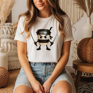 May include: White t-shirt with a cartoon coffee cup ninja design. The coffee cup has a black headband, a black band, and is holding two swords. The ninja coffee cup has an angry expression. The t-shirt is worn by a person.