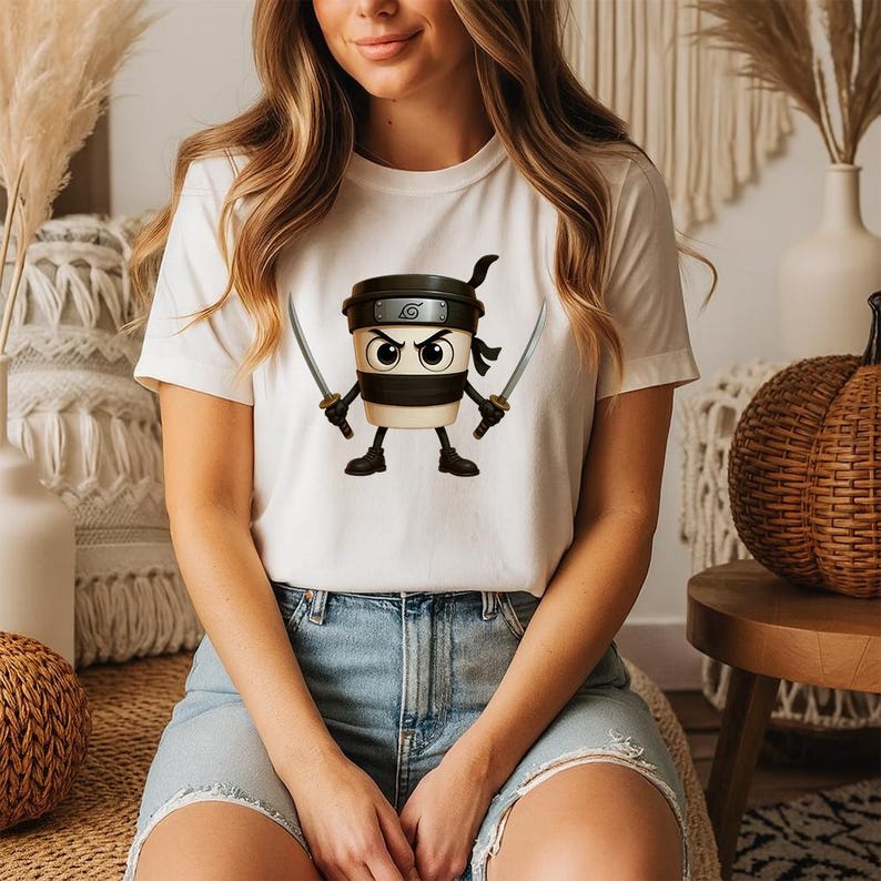 May include: White t-shirt with a cartoon coffee cup ninja design. The coffee cup has a black headband, a black band, and is holding two swords. The ninja coffee cup has an angry expression. The t-shirt is worn by a person.