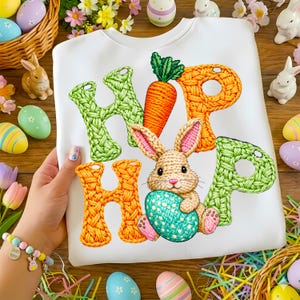 May include: White sweatshirt with a colorful "HOP" design featuring a carrot, a bunny holding an Easter egg, and green and orange letters. Easter-themed design with decorative eggs and bunnies.