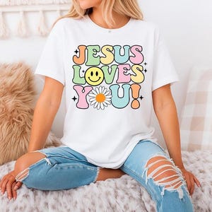 Könnte beinhalten: Weißes T-Shirt mit dem Text "JESUS LOVES YOU!" in farbenfrohen, Retro-Schriftzügen. Das Design beinhaltet ein Smiley, eine Gänseblümchen und Sternakzente. Die Person trägt zerrissene blaue Jeans.