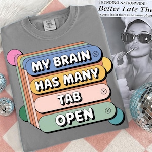 May include: Gray t-shirt with the text "MY BRAIN HAS MANY TAB OPEN" in a colorful, retro-style design. The graphic features stacked, rounded rectangles in blue, orange, pink, and green. A fun, casual tee.
