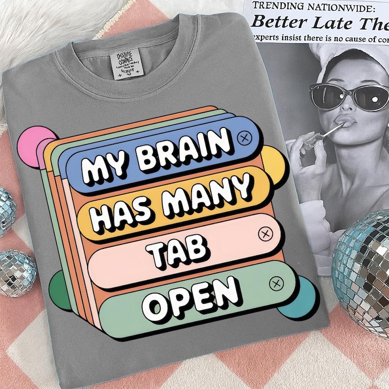 May include: Gray t-shirt with the text "MY BRAIN HAS MANY TAB OPEN" in a colorful, retro-style design. The graphic features stacked, rounded rectangles in blue, orange, pink, and green. A fun, casual tee.