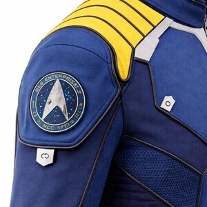 Puede incluir: Primer plano de una chaqueta de uniforme azul de Star Trek. La chaqueta presenta detalles amarillos en los hombros, un parche redondo con el logotipo de la USS Enterprise y detalles plateados. Un disfraz de ciencia ficci&oacute;n.