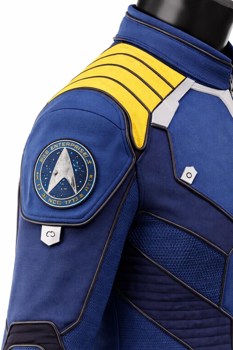 Puede incluir: Primer plano de una chaqueta de uniforme azul de Star Trek. La chaqueta presenta detalles amarillos en los hombros, un parche redondo con el logotipo de la USS Enterprise y detalles plateados. Un disfraz de ciencia ficci&oacute;n.
