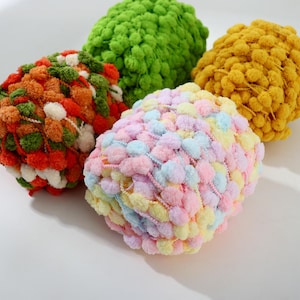 May include: Four balls of colorful pom-pom yarn in shades of green, orange, yellow, and pastel rainbow. The yarn is fluffy and textured, perfect for crafting projects like knitting or crocheting. The yarn is on a white background.