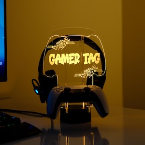 May include: A backlit gaming headset stand displaying the text "GAMER TAG". The stand, illuminated with a warm yellow light, holds a gaming controller and headset. The design features a stylized font.