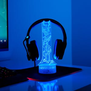 May include: A black gaming headset sits on a blue LED illuminated headphone stand. The stand has a circuit board design and is on a black mousepad. The background is a gradient of blue.