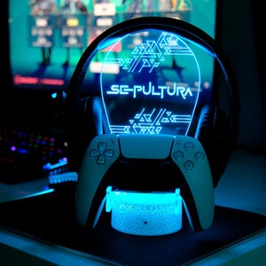 May include: A blue-lit gaming setup with a white controller, black headset, and a stand displaying the name "Sepultura". The stand and controller are white, and the headset is black. The setup is illuminated with blue light.