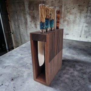 May include: A wooden knife block with a dark brown finish holds five knives with colorful handles. The block has a rectangular shape and is made of solid wood. The knives are arranged vertically in the block, with the handles facing up. The block is sitting on a gray surface.