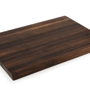 May include: A rectangular, dark brown wooden cutting board with a smooth surface. The board has a handle cut out on one side and a small logo in the corner. The wood grain is visible, and the board appears to be new.