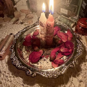 May include: Two lit candles, one white and one pink, stand upright on a silver-toned decorative tray. The tray is scattered with dried red rose petals and herbs. The candles have a textured, rustic appearance with visible botanical debris.