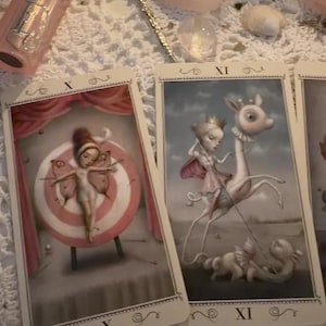 Puede incluir: Dos cartas de tarot marcadas con X y XI. La carta X muestra una figura con alas de mariposa sujeta a una diana rosa y blanca. La carta XI representa una figura coronada montando una criatura de cuello largo con una lanza hacia un dragón.