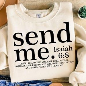 Puede incluir: Sudadera color crema con las palabras "send me" en letras negras grandes. Debajo está "Isaías 6:8" y una cita. Un sombrero beige y unos vaqueros azules son parcialmente visibles.