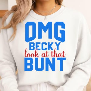 Può includere: Felpa bianca con la scritta "OMG BECKY look at that BUNT" in blu e rosso. Il testo è in grassetto. La felpa ha un girocollo e maniche lunghe.