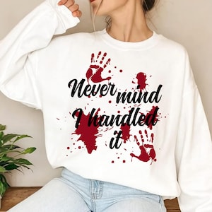 Puede incluir: Una sudadera blanca de manga larga con un diseño gráfico de huellas de manos rojas y salpicaduras similares a sangre. El texto negro en el centro dice "Never mind I handled it" escrito en una fuente cursiva sobre el área del pecho.
