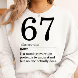 May include: White sweatshirt with the number "67" in large black font. Below the number is the word "noun" and a definition that reads "a number everyone pretends to understand but no one actually does."