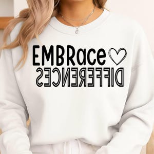 May include: White sweatshirt with the words "EMBRACE" and a heart symbol above the word "DIFFERENCE" in black block letters. The word "DIFFERENCE" is printed upside down. The sweatshirt is a crew neck style.