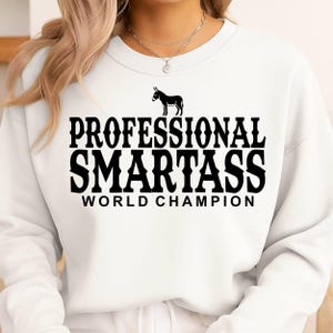 Puede incluir: Sudadera blanca con el texto "PROFESSIONAL SMARTASS WORLD CHAMPION" en letras negras. Un gráfico de burro negro está encima del texto. La sudadera es de estilo casual.
