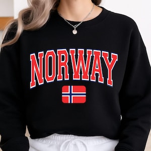 Puede incluir: Una sudadera negra de cuello redondo que presenta la palabra NORWAY en letras rojas grandes y arqueadas con un contorno blanco y azul. Debajo del texto hay un gráfico rectangular de la bandera de Noruega, con un fondo rojo y una cruz azul.