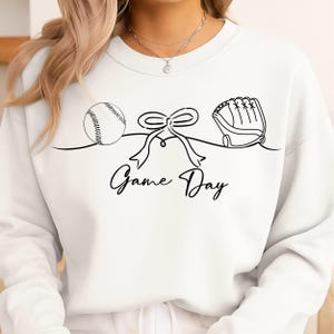Op de afbeelding: Witte sweatshirt met een zwarte lijntekening van een honkbal, een strik en een honkbalhandschoen. De woorden "Game Day" zijn in een scriptlettertype onder de tekening geschreven. De sweatshirt heeft een ronde hals.
