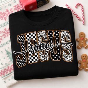 Könnte beinhalten: Ein schwarzes Sweatshirt mit Rundhalsausschnitt, das das Wort JESUS in großen Blockbuchstaben mit verschiedenen Mustern zeigt, darunter weiße Punkte, schwarz-weiße Karos, Leopardenmuster und weiße Sterne, mit dem Wort saves in weißer Schreibschrift.