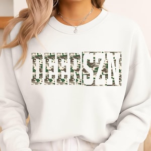 May include: A white crewneck sweatshirt featuring the text DEERSZN printed in a block font. The letters are filled with a camouflage pattern consisting of shades of green, brown, and beige. The sweatshirt has long sleeves and a ribbed neckline.