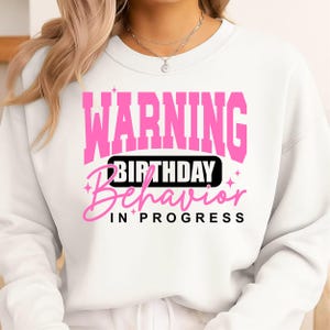 May include: White sweatshirt with the text "WARNING BIRTHDAY Behavior IN PROGRESS" in pink and black. The word "WARNING" is in a bold, blocky font, while "BIRTHDAY Behavior" is in a cursive font.