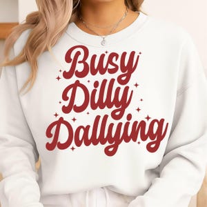 May include: White sweatshirt with the text "Busy Dilly Dallying" in a red, cursive font. The text is surrounded by small red starbursts. The sweatshirt has a crew neck and long sleeves.