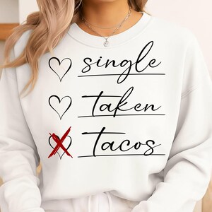 May include: White sweatshirt with the text "Single, Taken, Tacos" in black script. The word "Tacos" has a red X over the heart symbol. The sweatshirt has a crew neck and long sleeves.