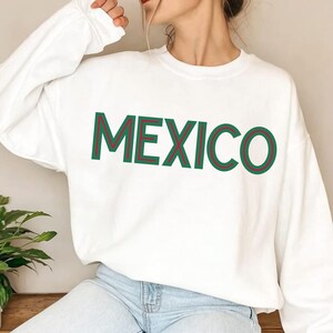 May include: A white long-sleeve crewneck sweatshirt featuring the word MEXICO printed in large capital letters across the chest. The lettering is styled in a dark green font with a thin red outline, creating a bold and clear graphic design.