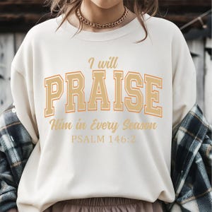 May include: Cream-colored sweatshirt with the text "I will PRAISE Him in Every Season" in a gold and orange collegiate font. The text "PSALM 146:2" is below. The sweatshirt is a casual style.