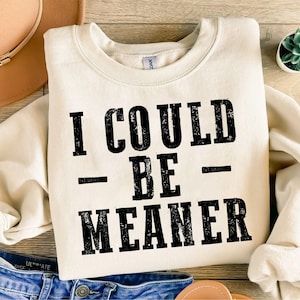 May include: Cream-colored sweatshirt with the text "I COULD BE MEANER" in bold, black letters. The sweatshirt is folded on a light wood surface, with a brown hat and blue jeans visible in the background. A small succulent plant is also present.