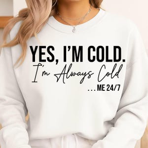 以下が含まれることがあります： 白のスウェットシャツ。黒い文字で「YES, I'M COLD. I'm Always Cold...ME 24/7」と書かれています。スウェットシャツはクルーネックと長袖です。シルバーのネックレスが見えます。