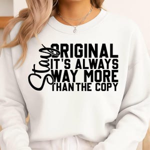 May include: White sweatshirt with the text "Stay Original It's Always Way More Than The Copy" in black. The sweatshirt has a crew neck and long sleeves. The text is in a bold, sans-serif font.