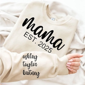 May include: A cream-colored sweatshirt with the word "Mama" in black script, and "EST. 2025" below. The sleeve displays the names "ashley", "taylor", and "britney" in black script. The sweatshirt is folded on a white surface.