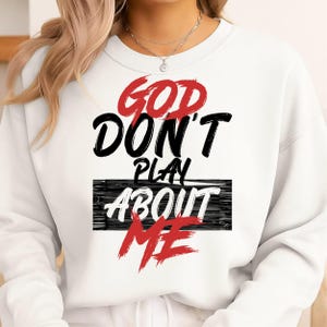 May include: White sweatshirt with the text "GOD DON'T PLAY ABOUT ME" in red, black, and white. The text is in a bold, stylized font. The sweatshirt is a crew neck style.