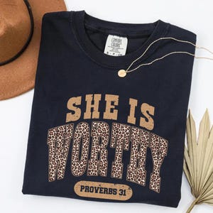 K&ouml;nnte beinhalten: Ein marineblaues T-Shirt mit dem Text "SHE IS WORTHY" in Braun und Leopardenmuster. Der Text "PROVERBS 31" steht darunter. Eine goldene Halskette und ein brauner Hut sind ebenfalls im Bild zu sehen.