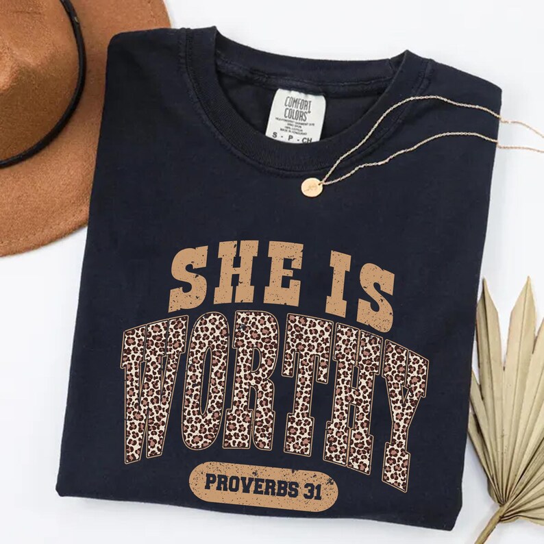 K&ouml;nnte beinhalten: Ein marineblaues T-Shirt mit dem Text "SHE IS WORTHY" in Braun und Leopardenmuster. Der Text "PROVERBS 31" steht darunter. Eine goldene Halskette und ein brauner Hut sind ebenfalls im Bild zu sehen.