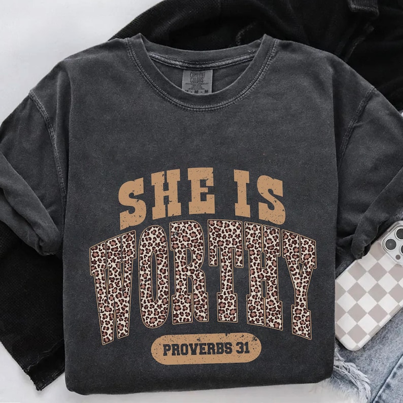 K&ouml;nnte beinhalten: Dunkelgraues Sweatshirt mit dem Text "SHE IS WORTHY" in Braun und Leopardenmuster. Der Text "PROVERBS 31" steht unter dem Wort "WORTHY". Das Sweatshirt ist gefaltet.