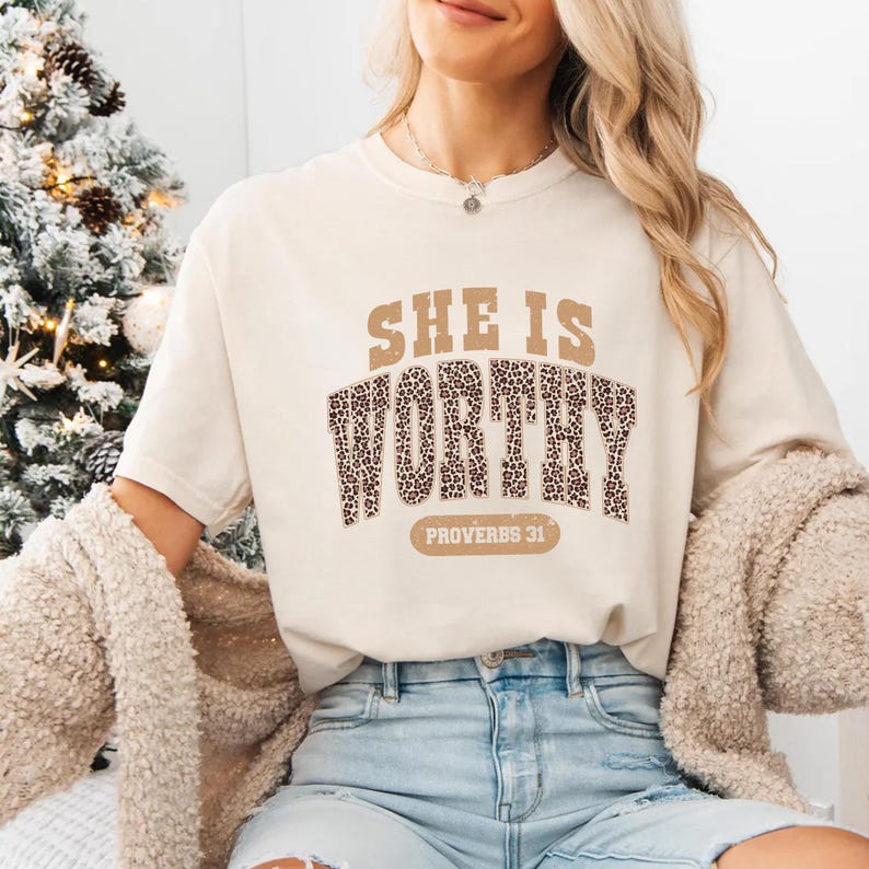 K&ouml;nnte beinhalten: Cremefarbenes T-Shirt mit dem Text "SHE IS WORTHY" in braunen und leopardenmuster Buchstaben. Der Text "PROVERBS 31" steht unter dem Wort "WORTHY". Das Shirt hat einen l&auml;ssigen Stil.