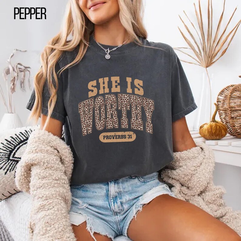 K&ouml;nnte beinhalten: Dunkelgraues T-Shirt mit dem Text "SHE IS WORTHY" in einem braunen und Leopardenmuster. Der Text "PROVERBS 31" steht darunter. Das T-Shirt hat einen l&auml;ssigen Stil.