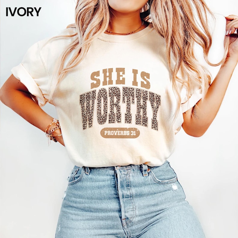 K&ouml;nnte beinhalten: Elfenbeinfarbenes T-Shirt mit dem Text "SHE IS WORTHY" in braunen Buchstaben mit Leopardenmuster. Darunter steht "PROVERBS 31". Das Shirt wird mit einer blauen Jeans kombiniert.