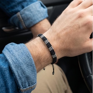 Könnte beinhalten: Ein gewebtes Armband in Schwarz und Grau wird am Handgelenk getragen. Das Armband hat ein geflochtenes Design mit abwechselnden schwarzen und grauen Abschnitten. Die Person trägt ein Jeanshemd mit hochgekrempelten Ärmeln.