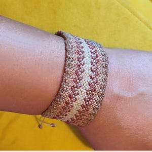 Könnte beinhalten: Ein breites gewebtes Armband mit einem sich wiederholenden Zickzackmuster in den Farben Beige, Creme und Rost. Das Textilband hat eine strukturierte Oberfläche mit sichtbaren Knoten und verfügt über dünne verstellbare Schnüre am unteren Rand.