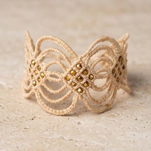Puede incluir: Una pulsera de macramé beige con un diseño calado detallado. La pulsera presenta una serie de bucles interconectados y está adornada con pequeñas cuentas doradas. El intrincado patrón y el color neutro la convierten en un accesorio versátil.