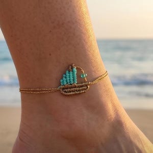 May include: Gold-colored anklet featuring a small boat design. The boat is crafted with turquoise beads and gold-toned thread, with a single turquoise bead accent. The anklet is worn on an ankle, with a beach and ocean background.