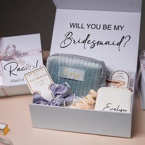 Könnte beinhalten: Eine weiße Geschenkbox mit dem Text "Will You Be My Bridesmaid?" enthält eine blau-grüne Kosmetiktasche, eine weiße Box mit dem Namen "Evelyn", ein Scrunchie und eine Karte. Zwei weitere Geschenkboxen sind sichtbar.