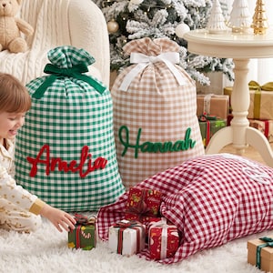 Pode incluir: Três grandes sacos de presente em tecido com padrão xadrez vichy em verde, bege e vermelho. O saco verde tem o nome Amelia em vermelho, o bege tem o nome Hannah em verde, e ambos estão atados com uma fita. Pequenos presentes embrulhados por perto.