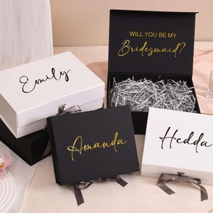 以下が含まれることがあります： 白と黒のギフトボックスのコレクション。 「Emily」、「Amanda」、「Hedda」などの名前がエレガントな筆記体で書かれています。 1つのボックスは開いており、細断された紙と「Will You Be My Bridesmaid?」というテキストが金色で表示されています。 各ボックスにはリボンが付いています。